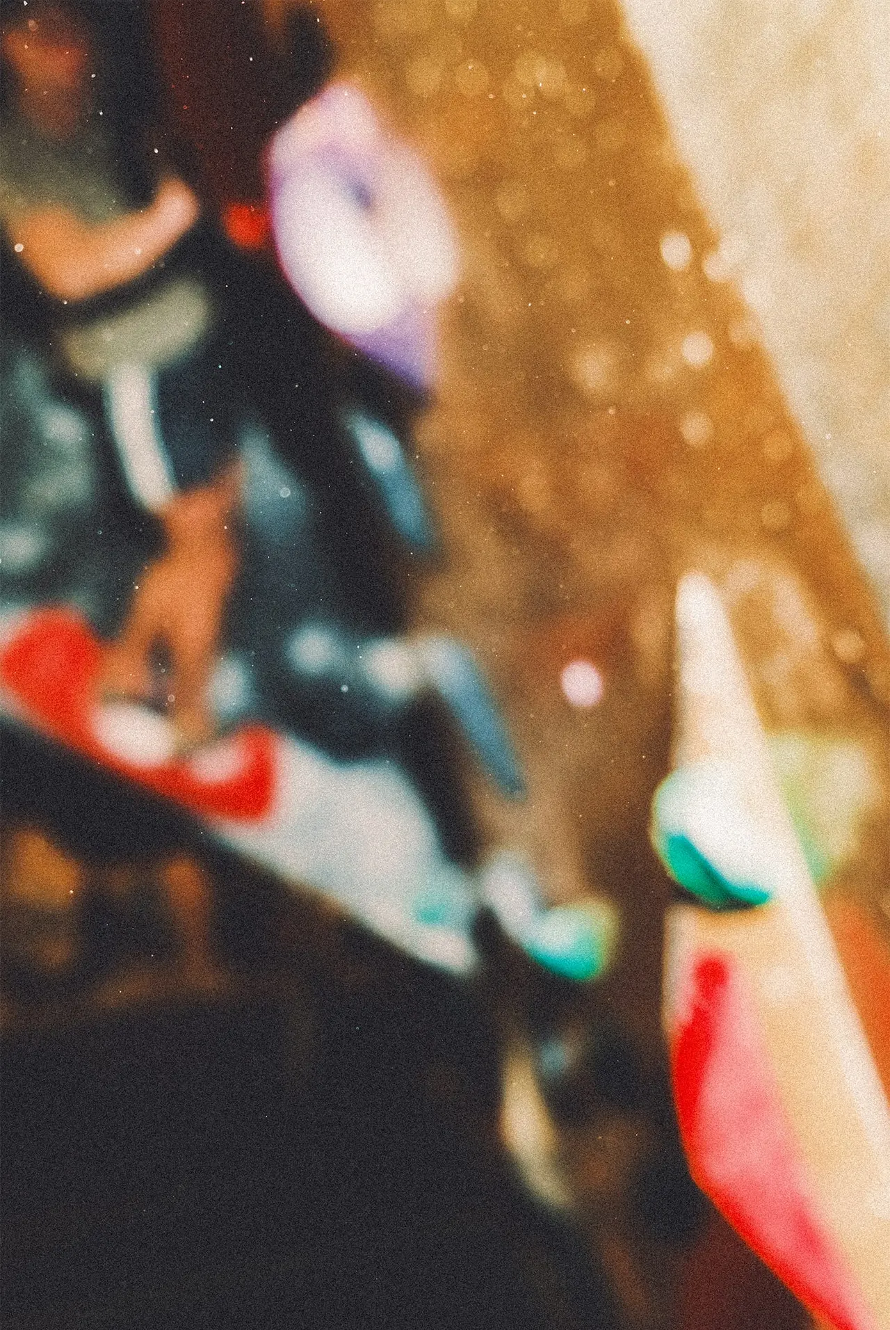 Heavily blurred close-up of climbing holds in red, green and turquoise on a tan-coloured wall with climbers in background.