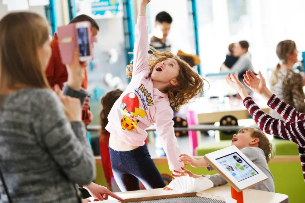 Children engaged in an interactive activity at an indoor science centre, with one child reaching upward excitedly.
