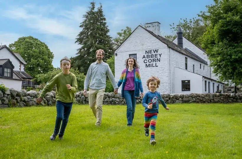 Family of four running across a grass field towards a white stone mill building on a sunny day.
