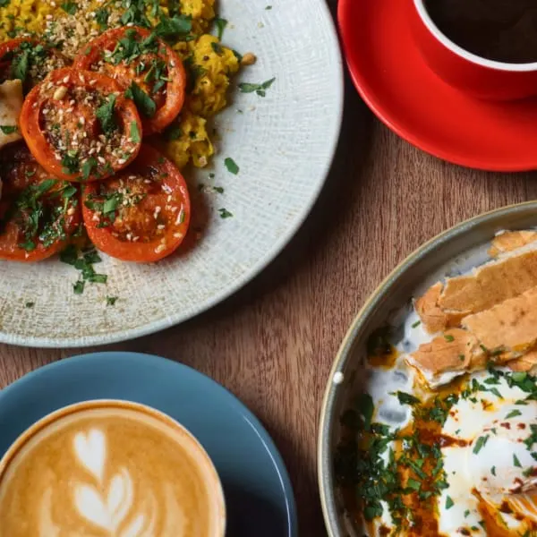 Overhead view of a café table with tomato dish, coffee with latte art, pastry, and red cup on wooden surface.
