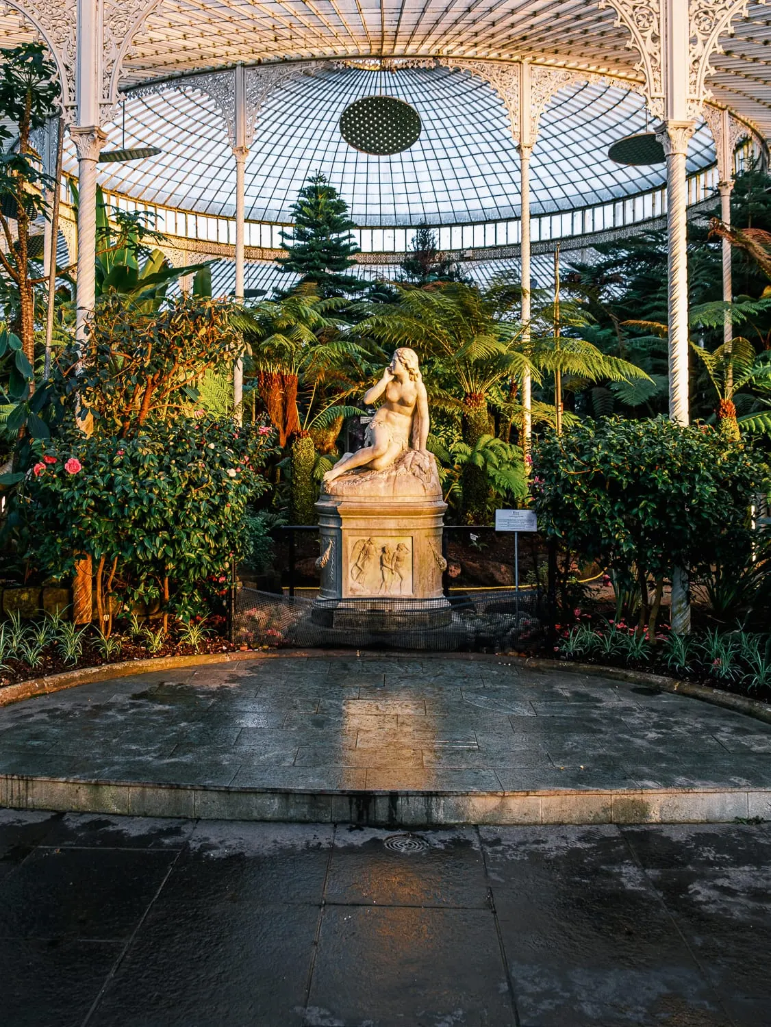 Classical statue on a pedestal surrounded by tropical plants inside an ornate glass and cast-iron greenhouse structure.