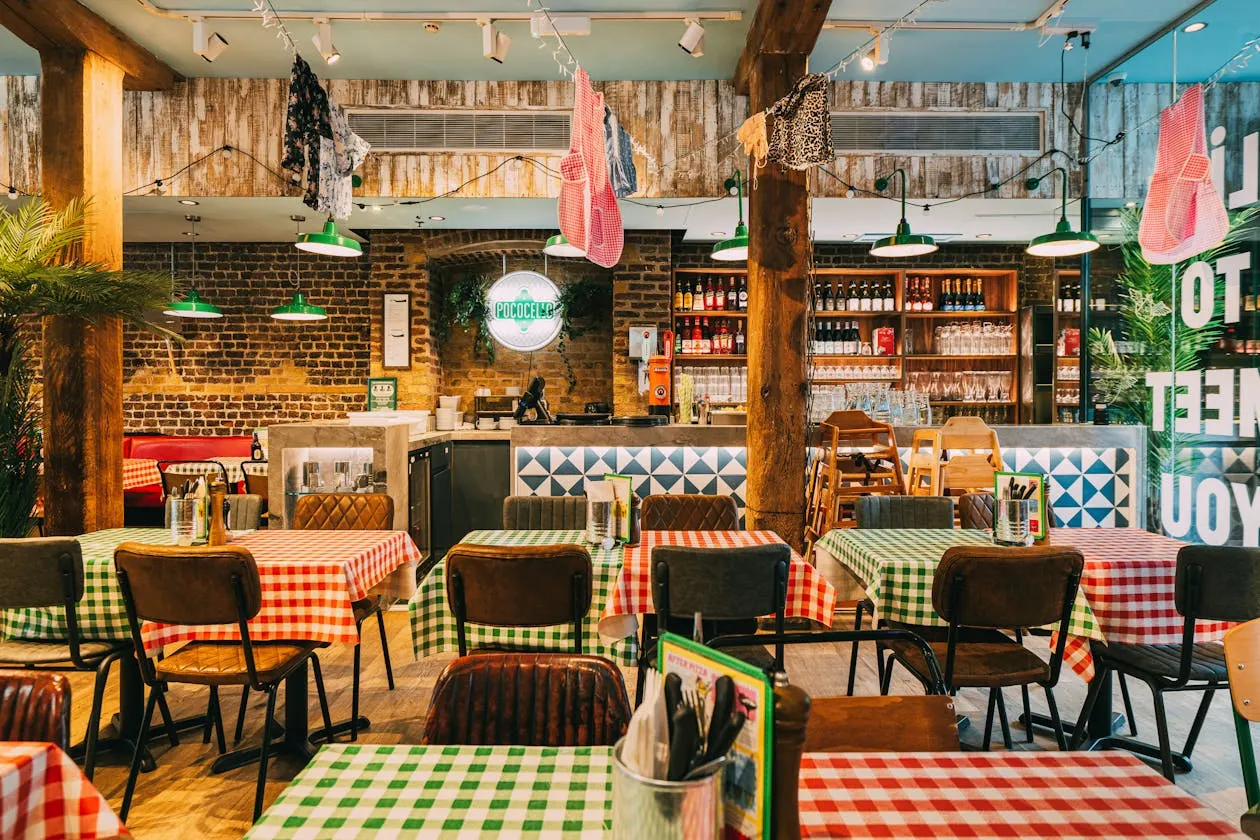 A casual restaurant interior with red and white gingham tablecloths, exposed brick walls, hanging clothes, green pendant lights, wooden beams, and a service counter.