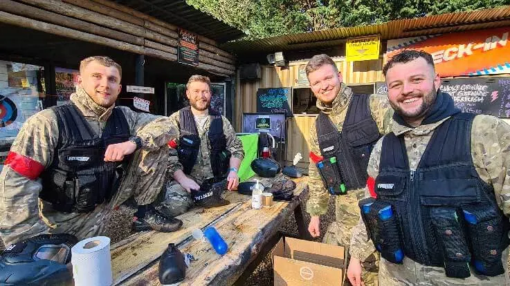 Four men in camouflage clothing and tactical vests stand smiling at a wooden table inside a covered outdoor shelter at an adventure park.