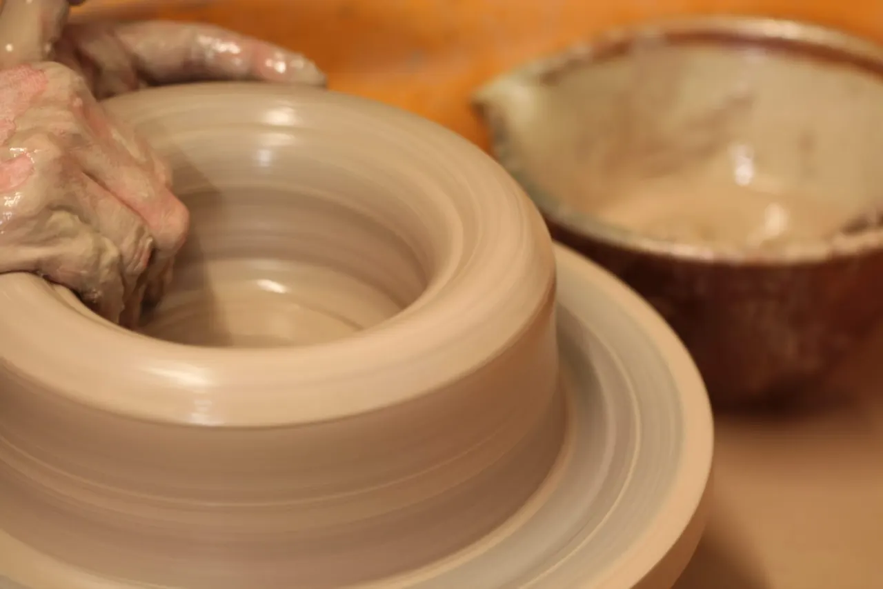 Potter's hands shaping wet clay on a spinning wheel, with finished vessels blurred in the background.