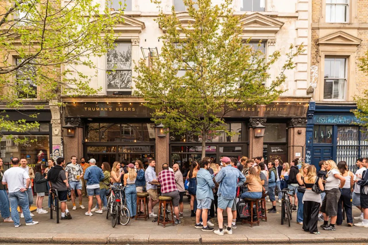Busy outdoor street scene with crowds of people gathered outside a pub, some seated on stools with drinks, trees overhead, bicycles parked nearby.