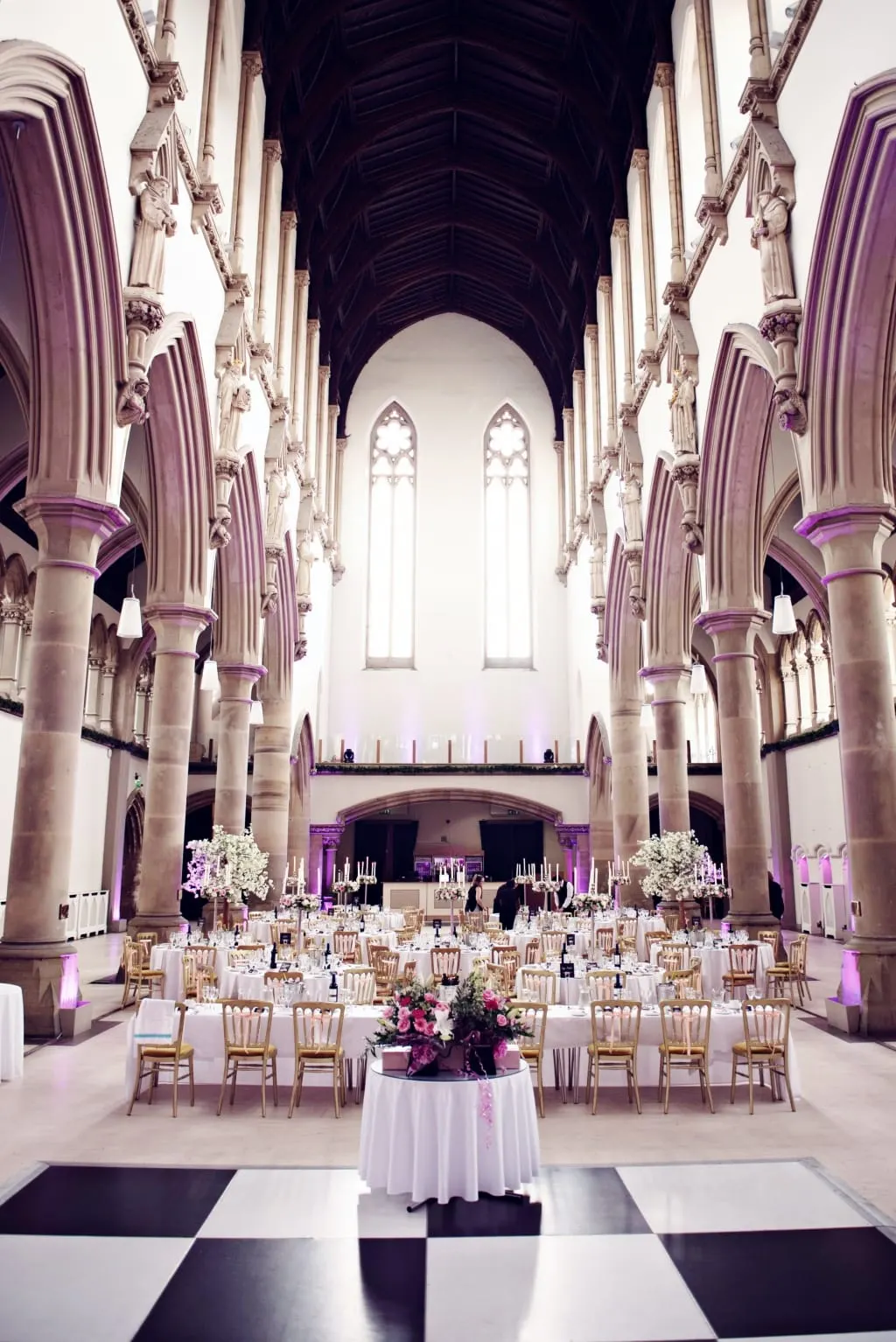 Gothic cathedral interior set up for a formal event with rows of tables, chairs, floral arrangements and purple lighting beneath soaring stone columns and arched windows.