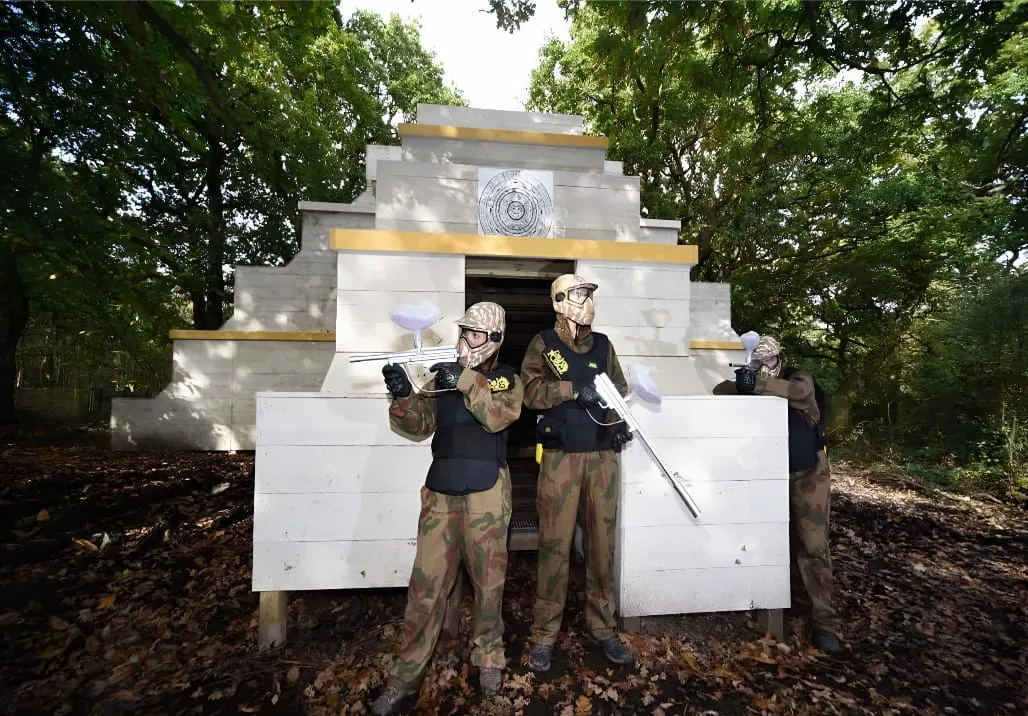 Players in camouflage and protective gear holding paintball markers in front of a white wooden fort structure in a wooded area.