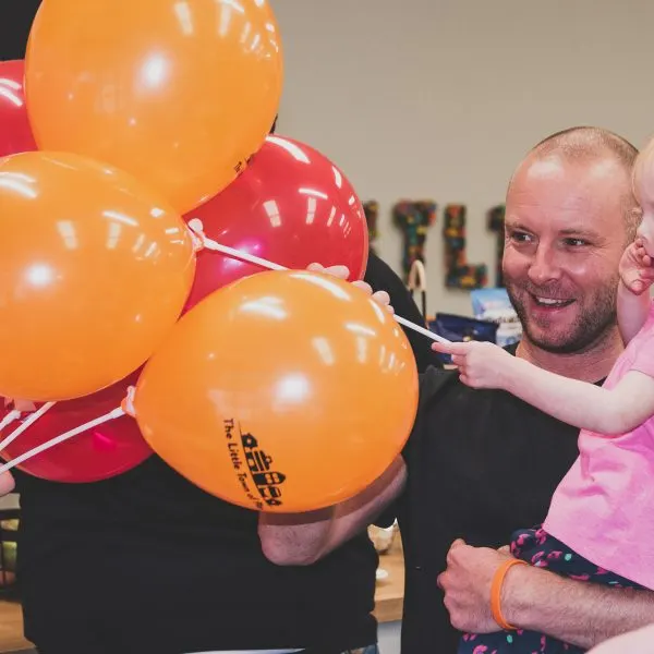Man holding a young child surrounded by orange and red balloons at an indoor play venue.