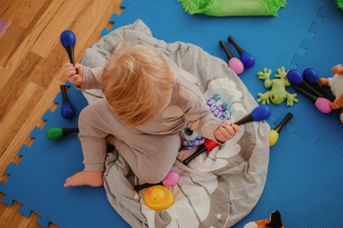 Toddler playing with colourful toys and mallets on a padded blue mat in an indoor play space.