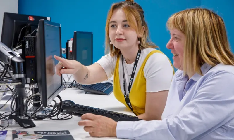 Two women at a computer workstation, one wearing a yellow lanyard, pointing at a monitor screen.