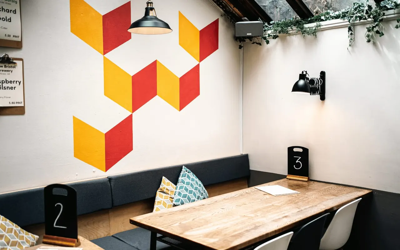Interior of a board game café with a wooden table, navy booth seating, geometric wall mural in yellow and red, and industrial pendant lights.