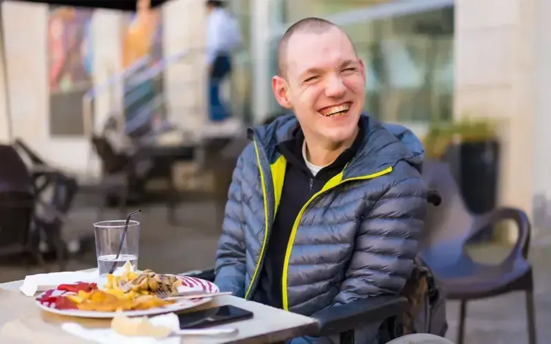 Young person in navy padded jacket smiling at table with plate of food and glass of water indoors.