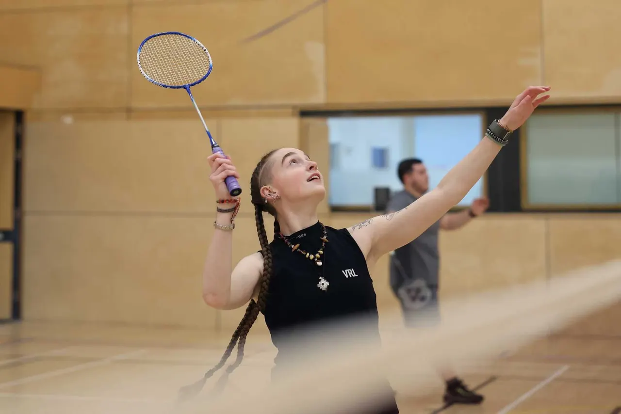 Young woman playing badminton indoors, holding a blue racket with arm raised, in a sports hall with other players in the background.