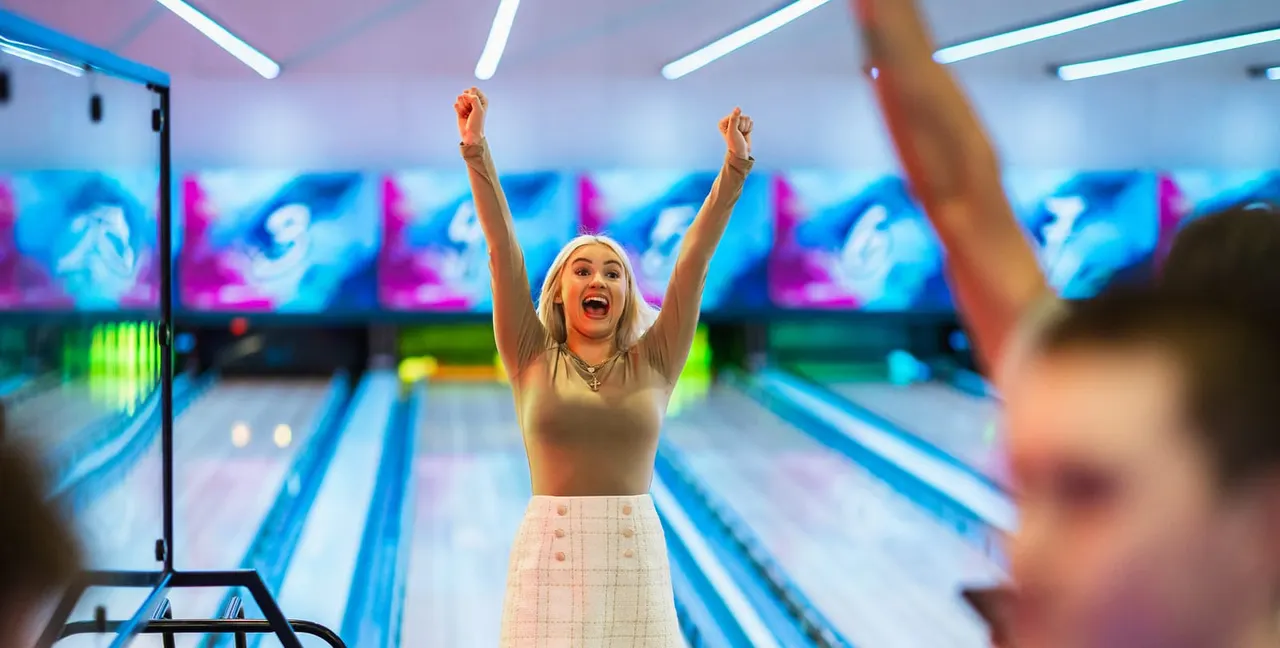Woman celebrating with arms raised at a bowling alley with illuminated lanes and digital scoring displays.