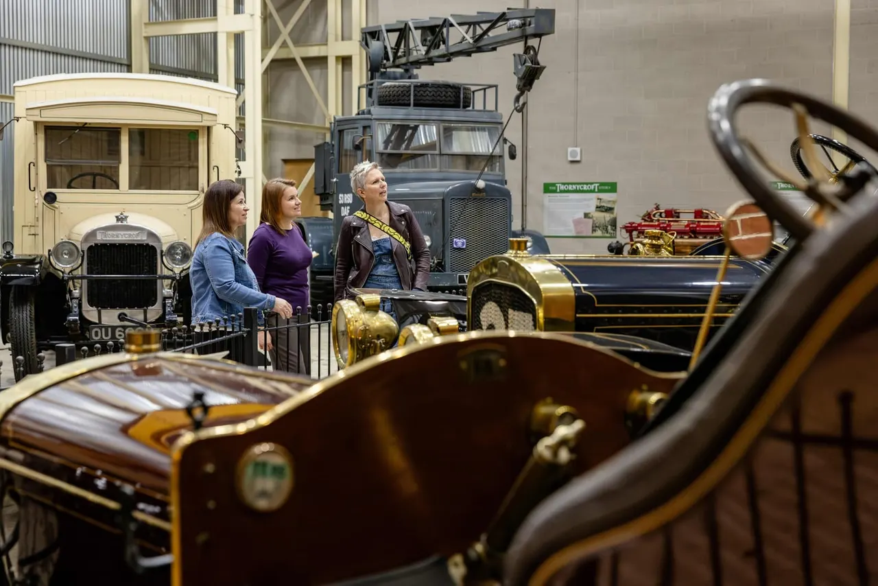 Three visitors admire a collection of vintage and classic vehicles displayed in an indoor museum hall.