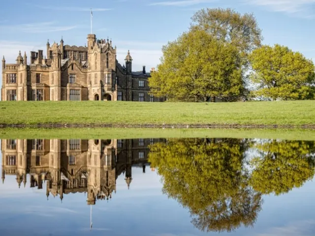 Stone castle with towers reflected in still water, with green field and trees beyond.