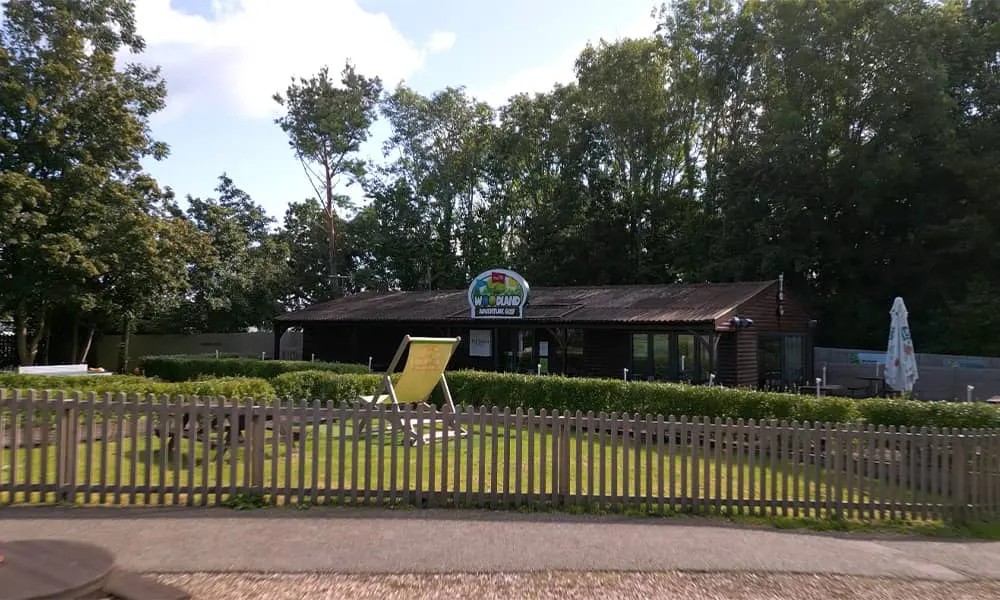 A low wooden leisure building with yellow picket fencing, a large deckchair and signage visible, surrounded by mature trees on a sunny day.