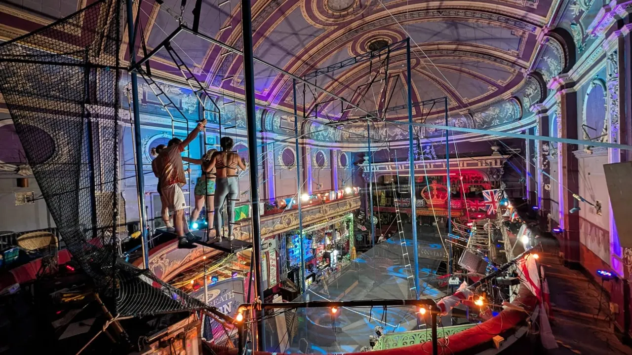 Children on a trapeze with safety net inside an ornate circus ring with purple and gold decorated ceiling and atmospheric lighting.