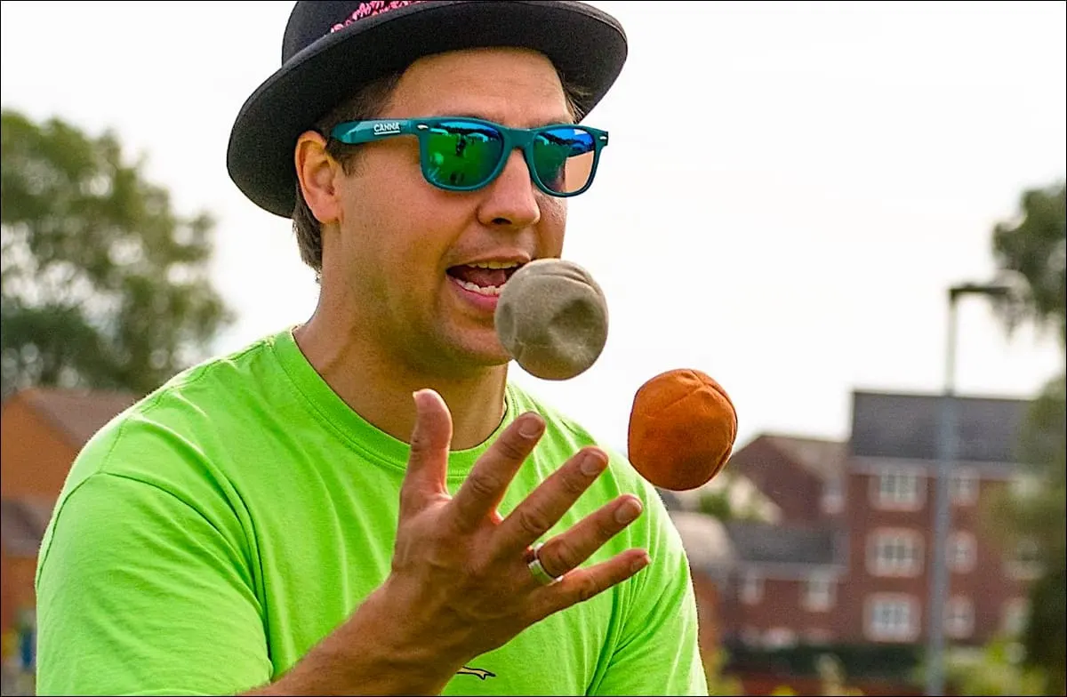 Man in bright green shirt and sunglasses juggling balls outdoors with buildings in background.