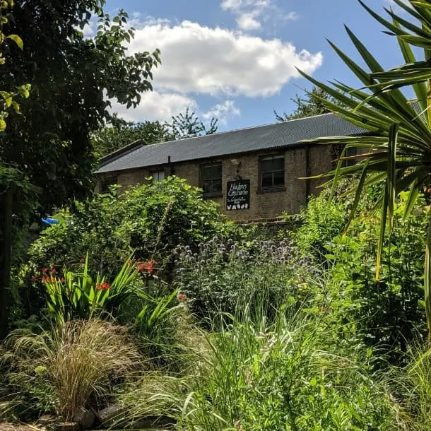 Stone building with signage surrounded by lush green plants and ornamental grasses under blue sky.