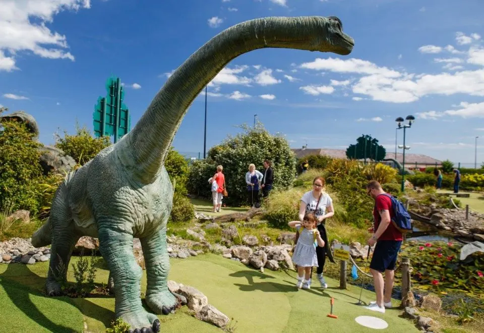 Families playing mini golf around a large dinosaur sculpture in a themed outdoor course.