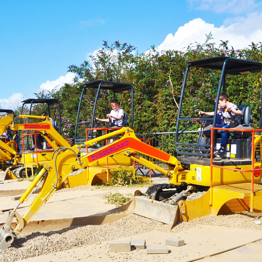 Young children operating toy diggers and excavators in a supervised play area on a sunny day.