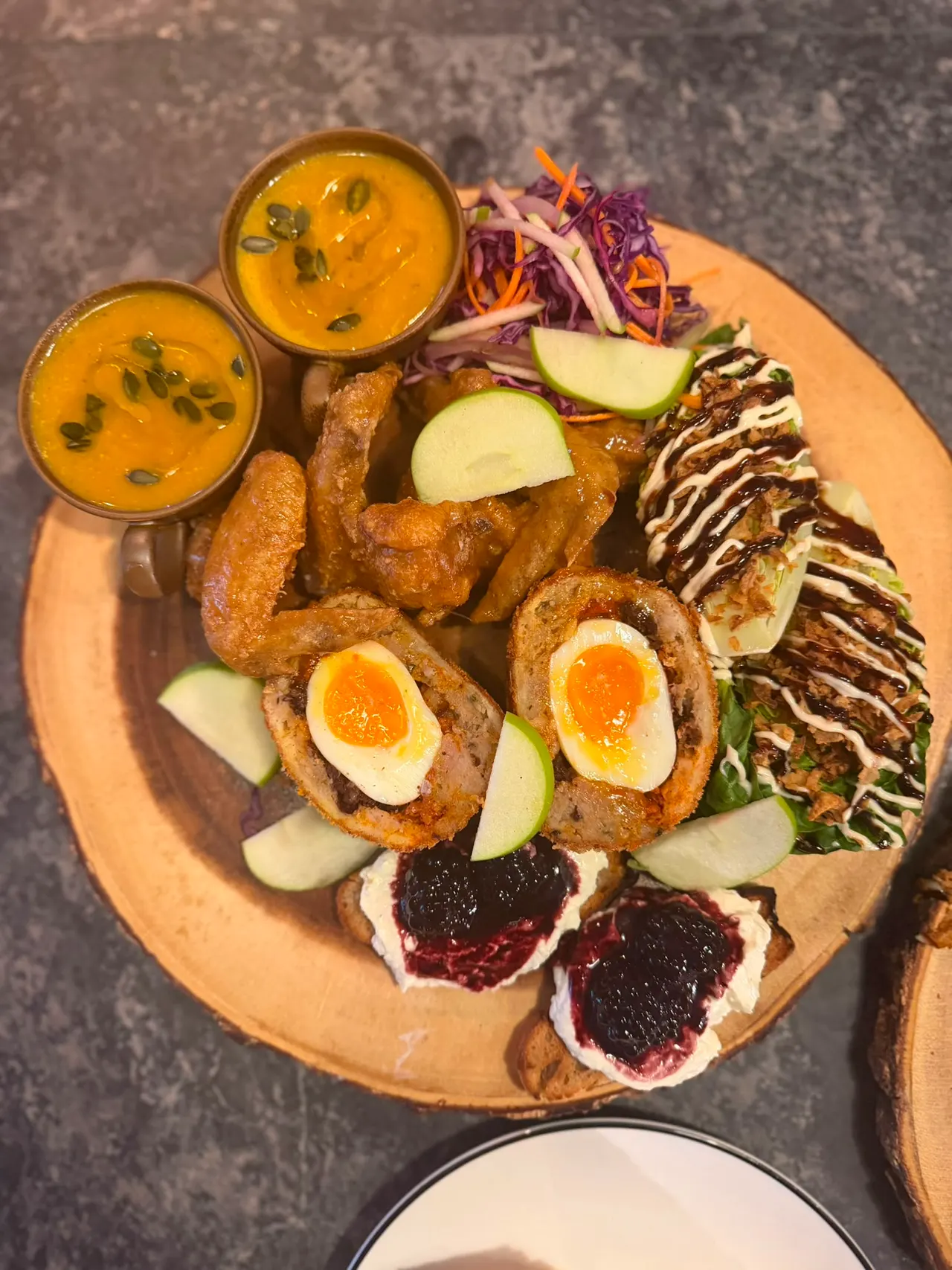 A wooden board displaying an assortment of cured meats, soft cheese, boiled eggs, apple slices, shredded vegetables and two small bowls of golden sauce.