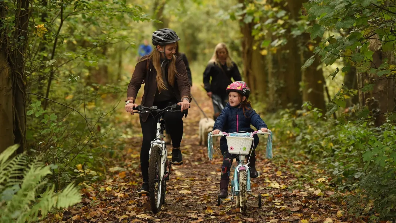 Woman and child cycling on a tree-lined woodland path with another person walking behind.