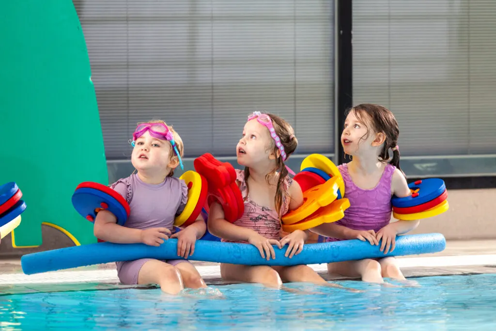 Three young children in swimming costumes and colourful swim aids sit together in a swimming pool holding a blue foam noodle.