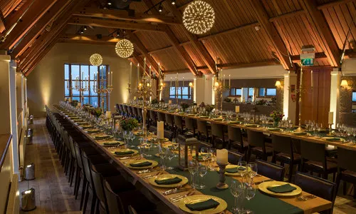 Long dining table set for formal event in wood-beamed hall with decorative pendant lights and evening views through windows.