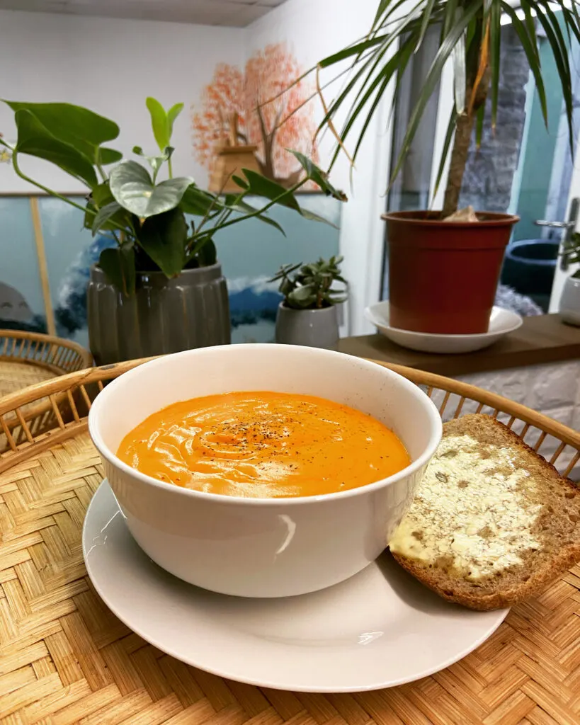 Bowl of orange soup with bread roll on woven placemat, potted plants behind on table indoors.