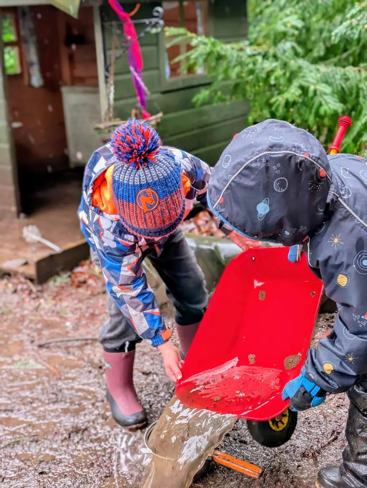 Two young children in winter clothing and hats play in a muddy puddle with a red toy truck outdoors.