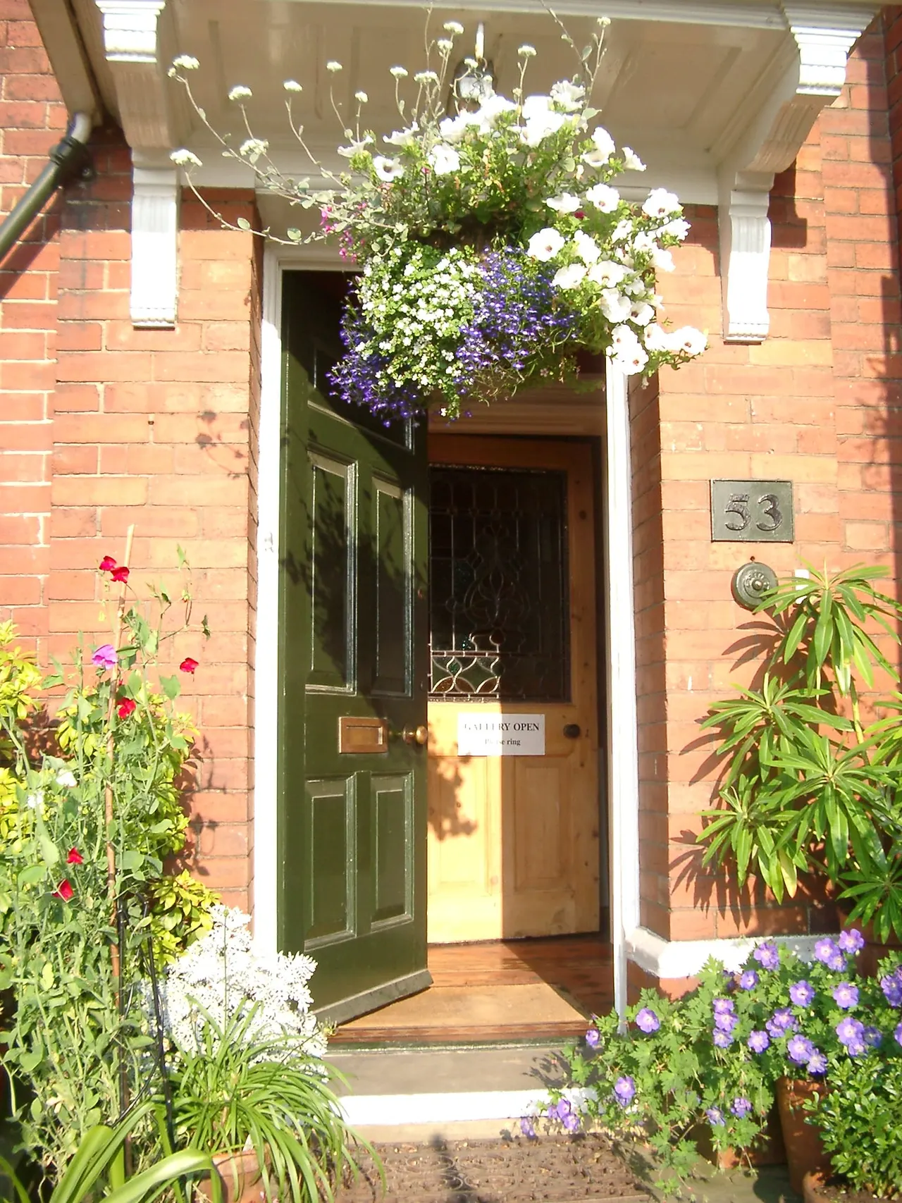 Entrance to a brick building with a green door framed by white columns, decorated with hanging baskets and potted flowers in white, purple and red.