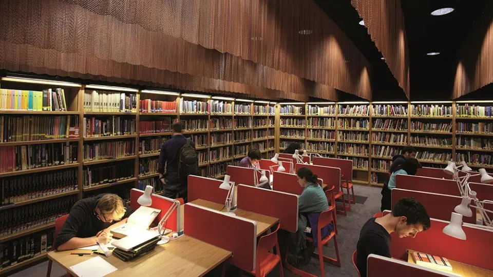 Several visitors study at individual desks with red partitions in a modern library reading area lined with bookshelves.