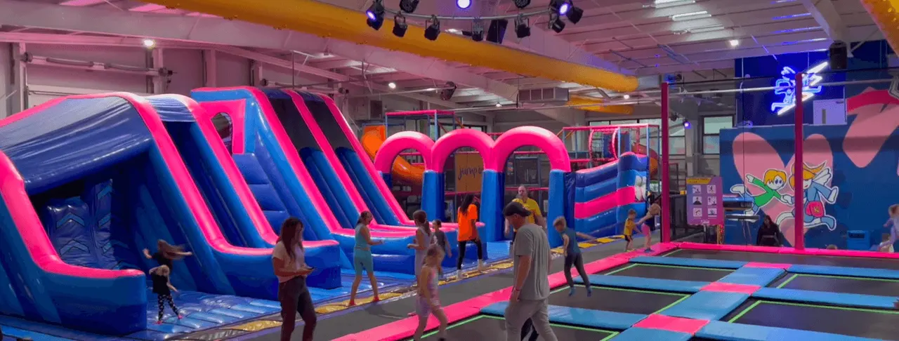 Children and adults playing on inflatable slides and trampolines in a brightly lit indoor leisure facility.