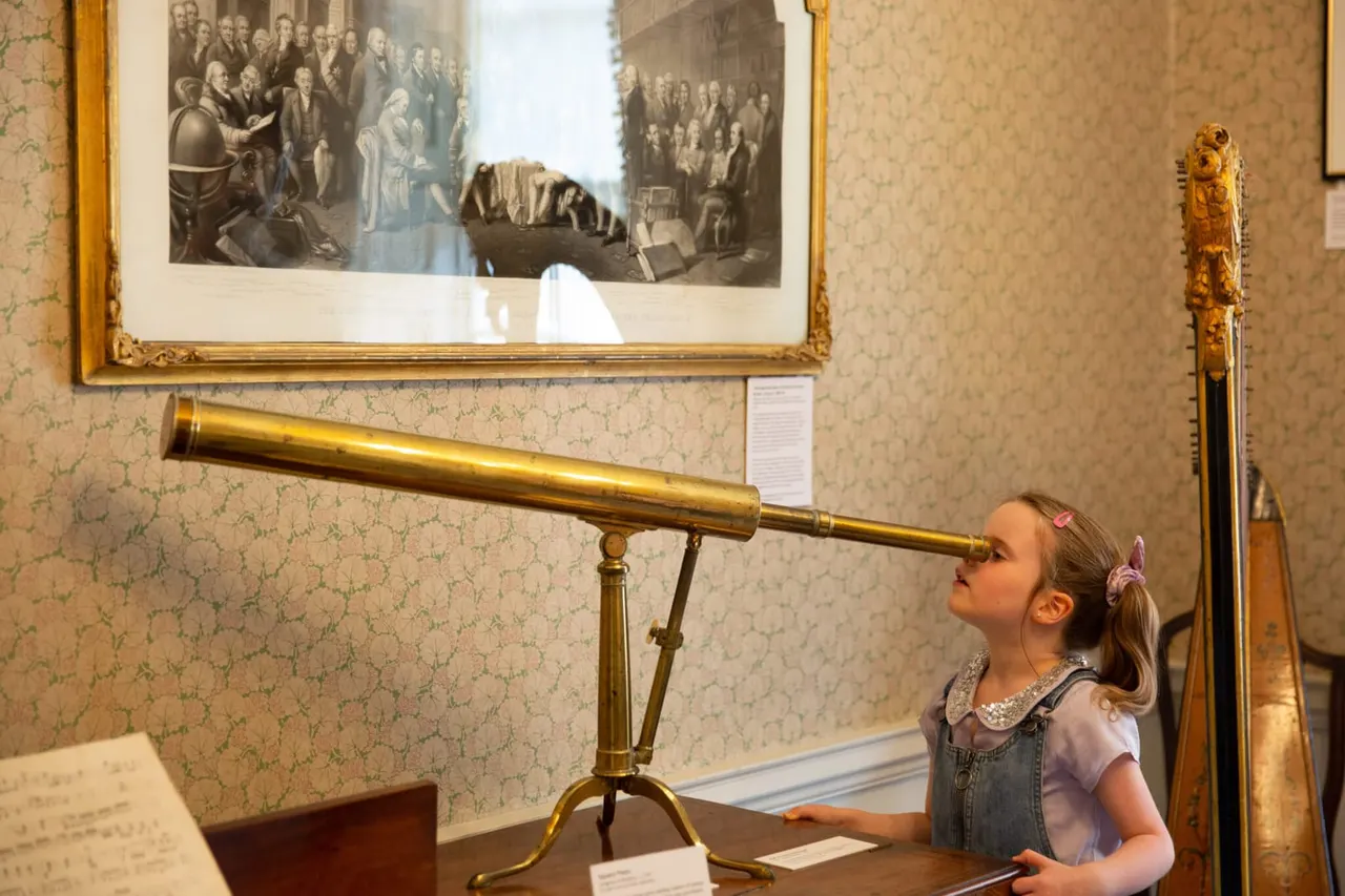 Young child looking through a brass telescope in a museum room with historical engravings and wallpapered walls.