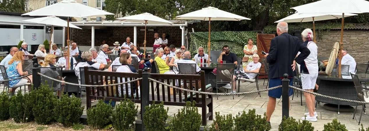Large group of older adults socialising under white umbrellas on a paved outdoor bowling club terrace with wooden benches and hedges.