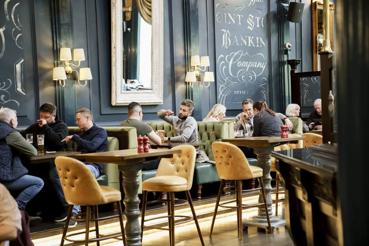 Groups of people seated at tables and in booth seating throughout an elegant pub interior with dark walls, gold sconces and ornate signage.