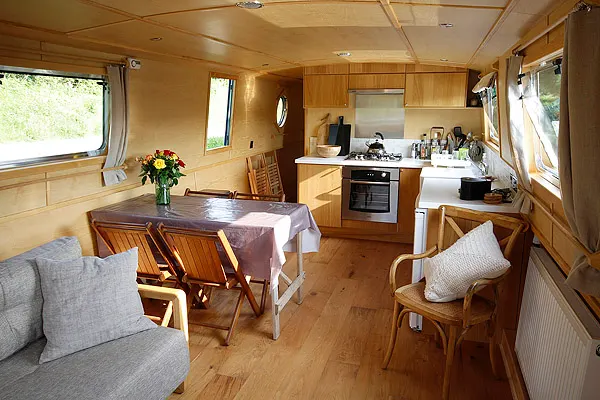 Interior of a wooden narrowboat showing dining table, kitchen area, seating and windows.