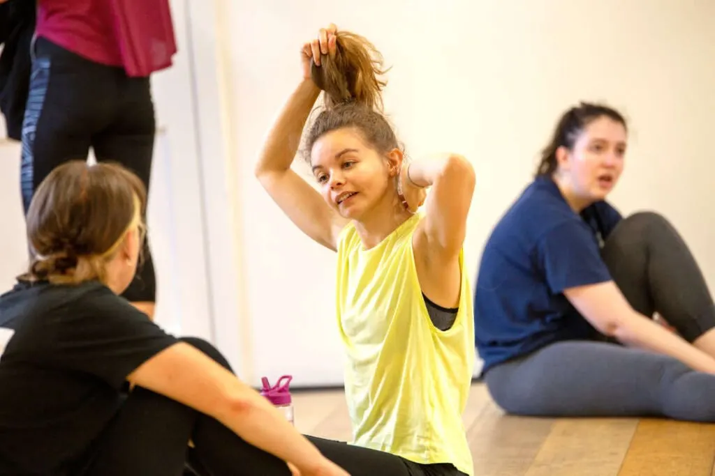 Girl in yellow tank top adjusting her hair during a dance class with other participants sitting on the floor.