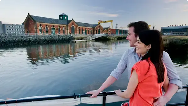 A man and woman on a boat tour on a waterway, with a historic red-brick building and crane visible across the water.