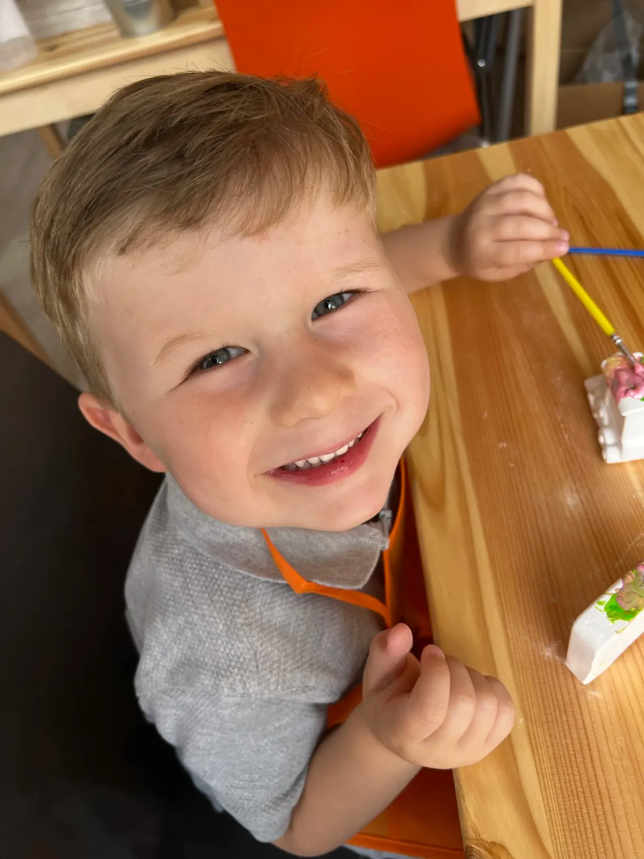 Young child smiling at a table whilst holding a paintbrush during an activity session.