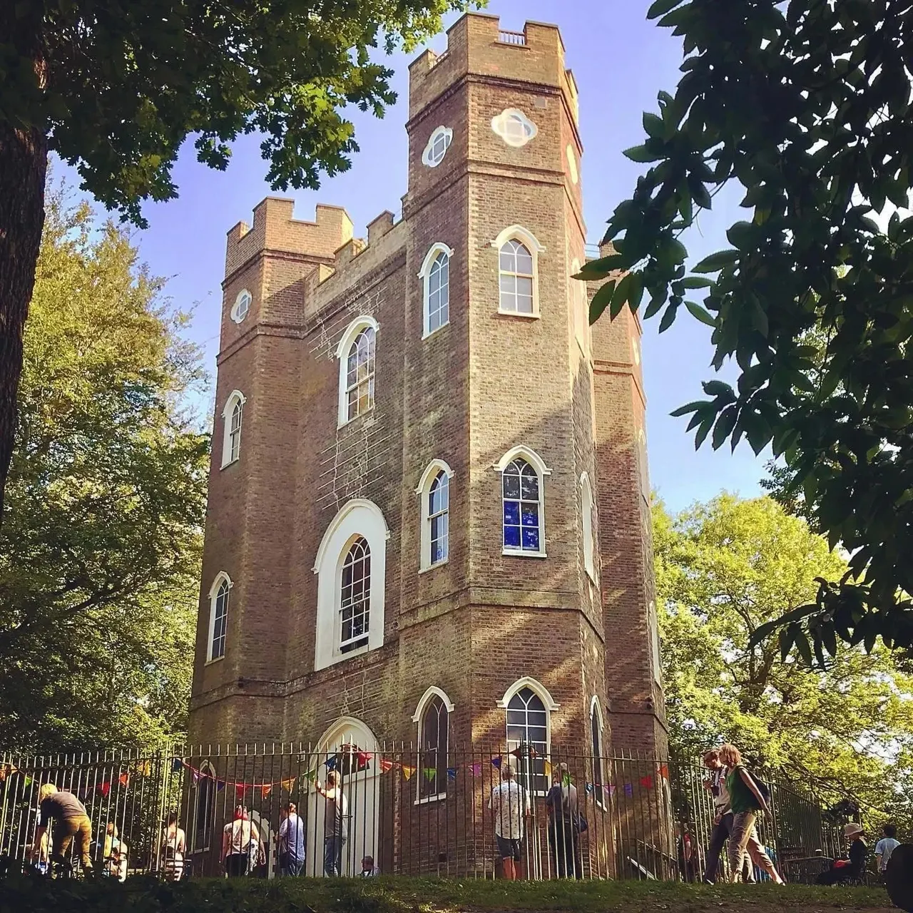 Historic brick tower with crenellated battlements surrounded by trees and visitors gathered at its base on a sunny day.