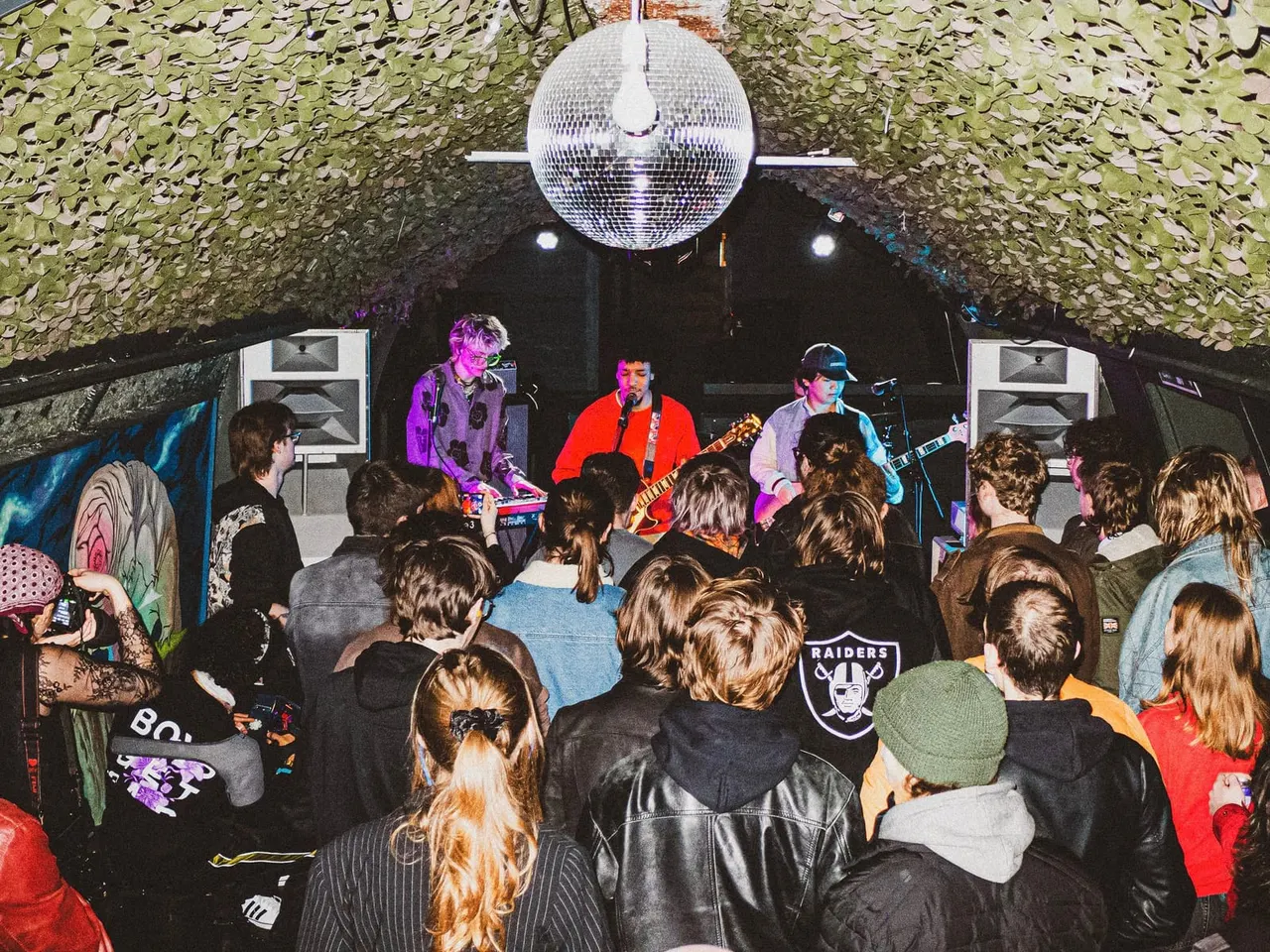 A live band performs on stage under a glittering disco ball to a crowd of young people in an indoor venue with green foliage decoration.