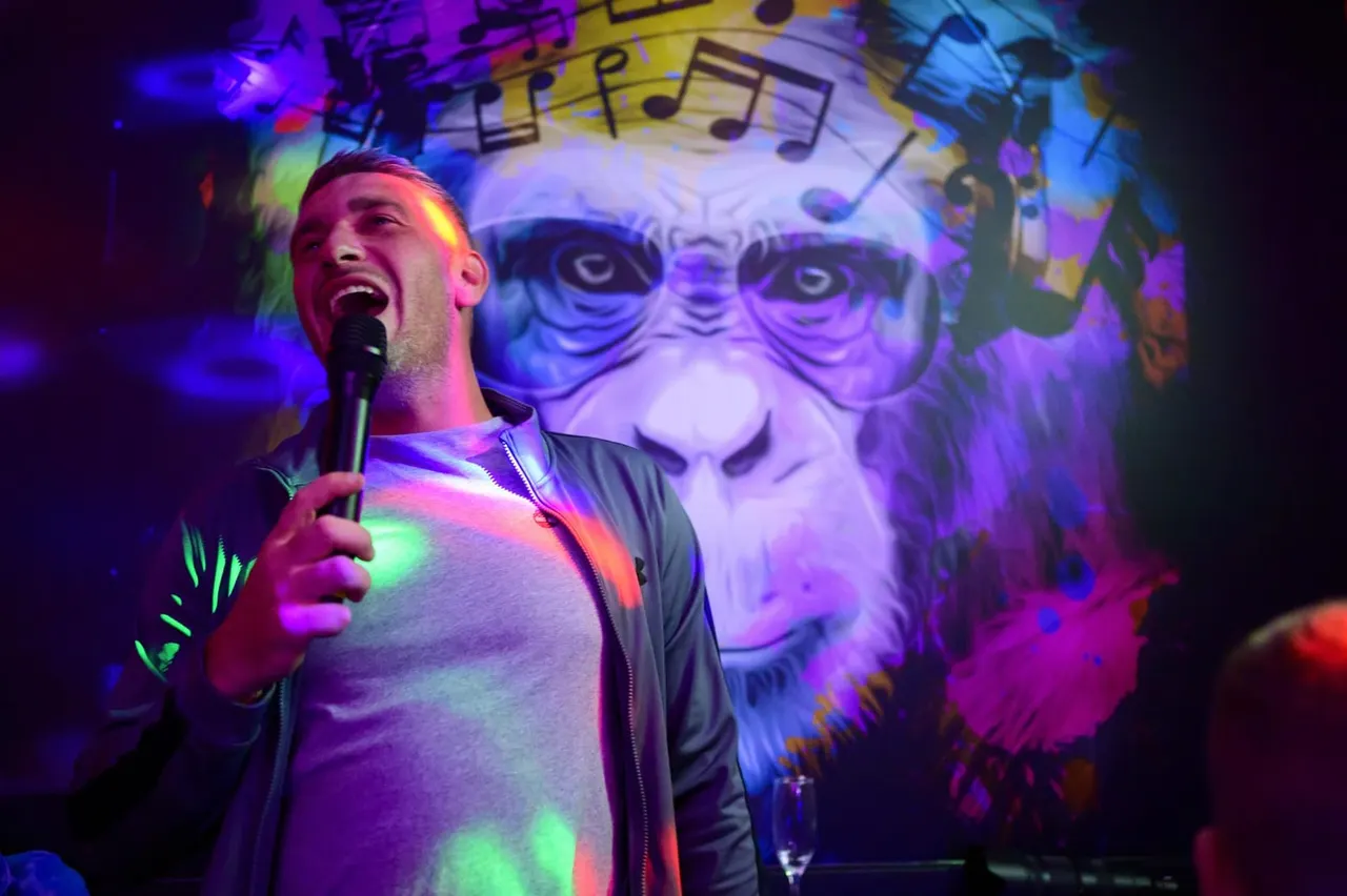 Man singing into a microphone on stage with colourful lighting and musical note graphics projected behind him.