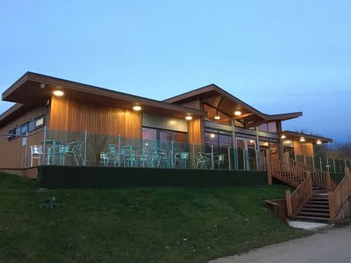 Modern wooden leisure venue with lit interior, deck seating and stairs, set on grassy hillside at dusk.