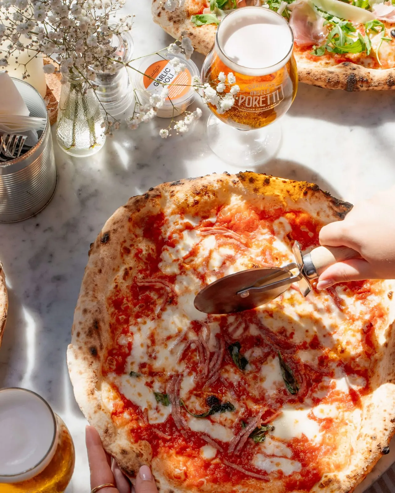 Person cutting into wood-fired pizza with tomato sauce and cheese, served with beer and flowers on marble table.
