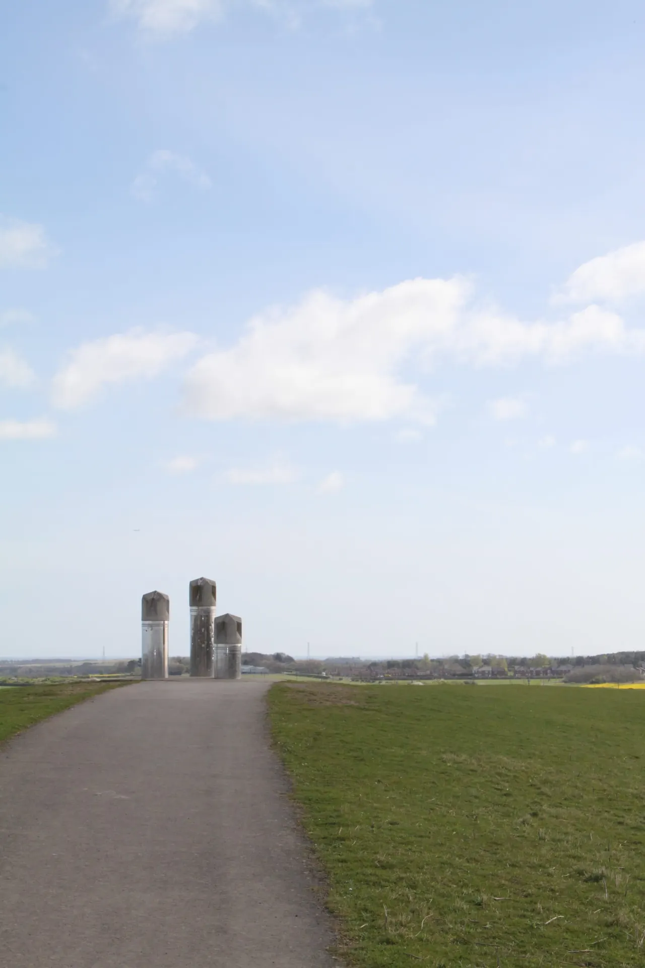 Paved pathway through open grassland leading towards three cylindrical structures under a blue sky with white clouds.