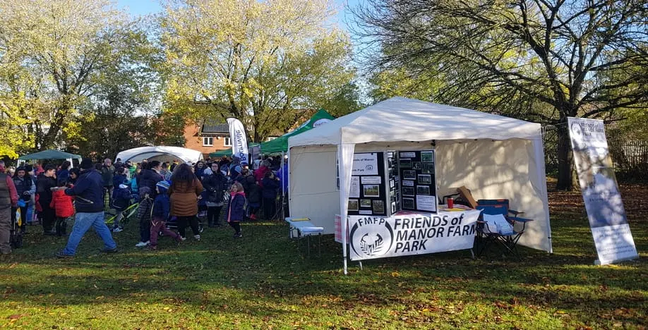 Outdoor community event with visitors gathering around information tents and marquees in a leafy park setting.