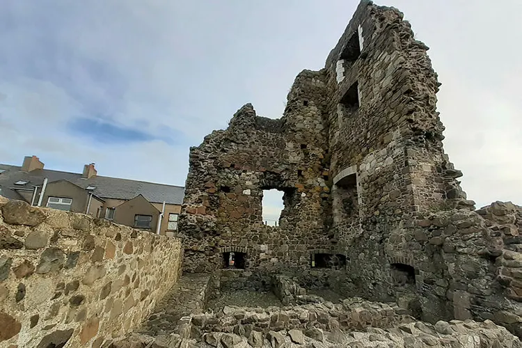 Ruined stone castle tower with window openings, adjacent to modern residential building under cloudy sky.